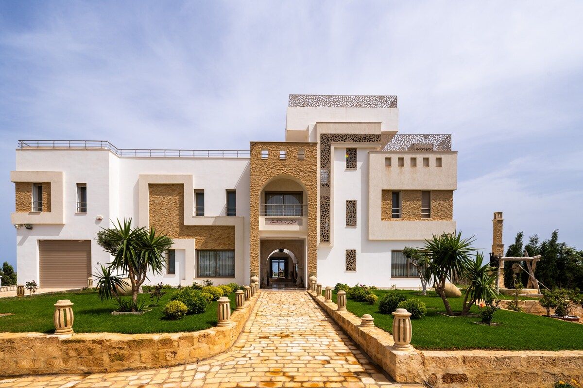 Façade principale de la villa Dar Aïcha à Tazarka, architecture blanche contemporaine avec pierre locale et jardin méditerranéen
